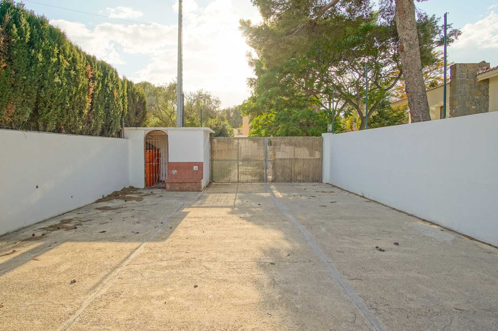 Entrada de la propiedad con amplio acceso pavimentado, muros perimetrales blancos y puerta metálica con ocultación, rodeada de pinos y setos de ciprés.