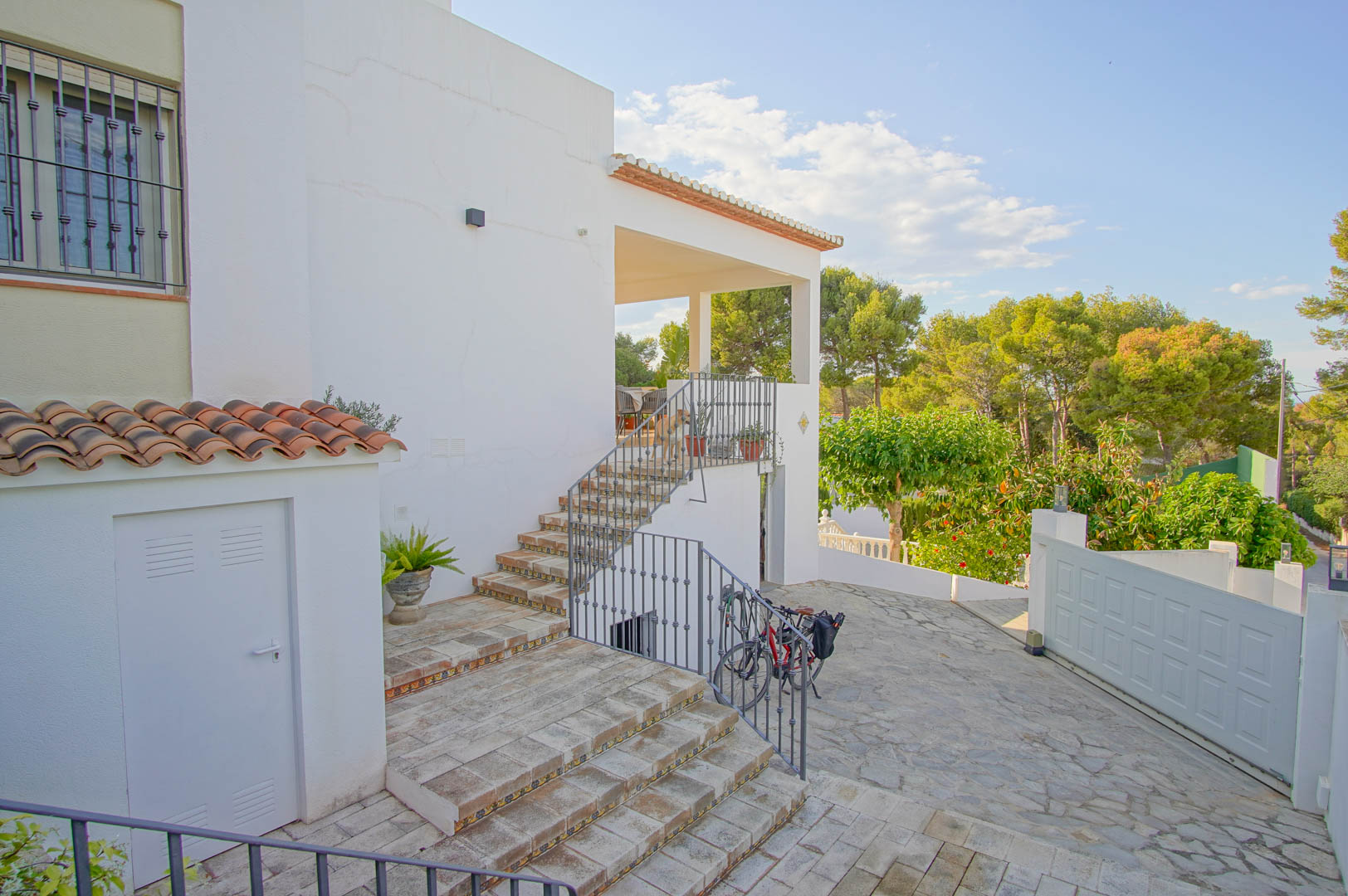 Entrada exterior con escaleras de gres y detalles cerámicos que conducen a la terraza cubierta, rodeada de vegetación mediterránea y pinos.