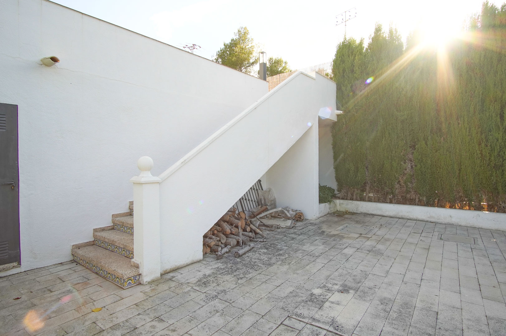 Escalera exterior de obra con peldaños de piedra y azulejos decorativos, situada junto a un patio pavimentado y zona de almacenaje de leña.