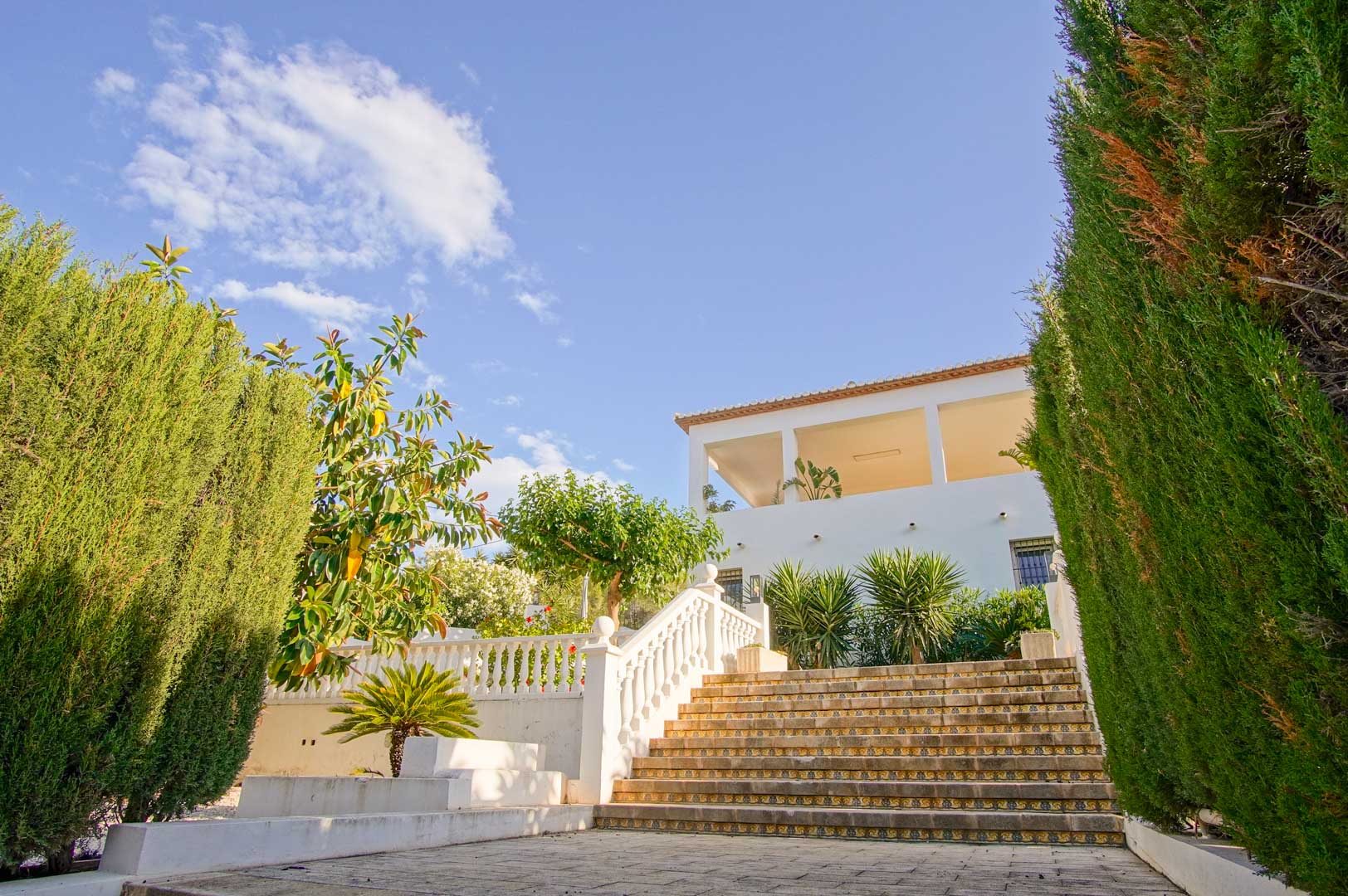 Escalera exterior de piedra con azulejos decorativos y balaustrada blanca, flanqueada por altos cipreses que conducen a la fachada principal de la villa.