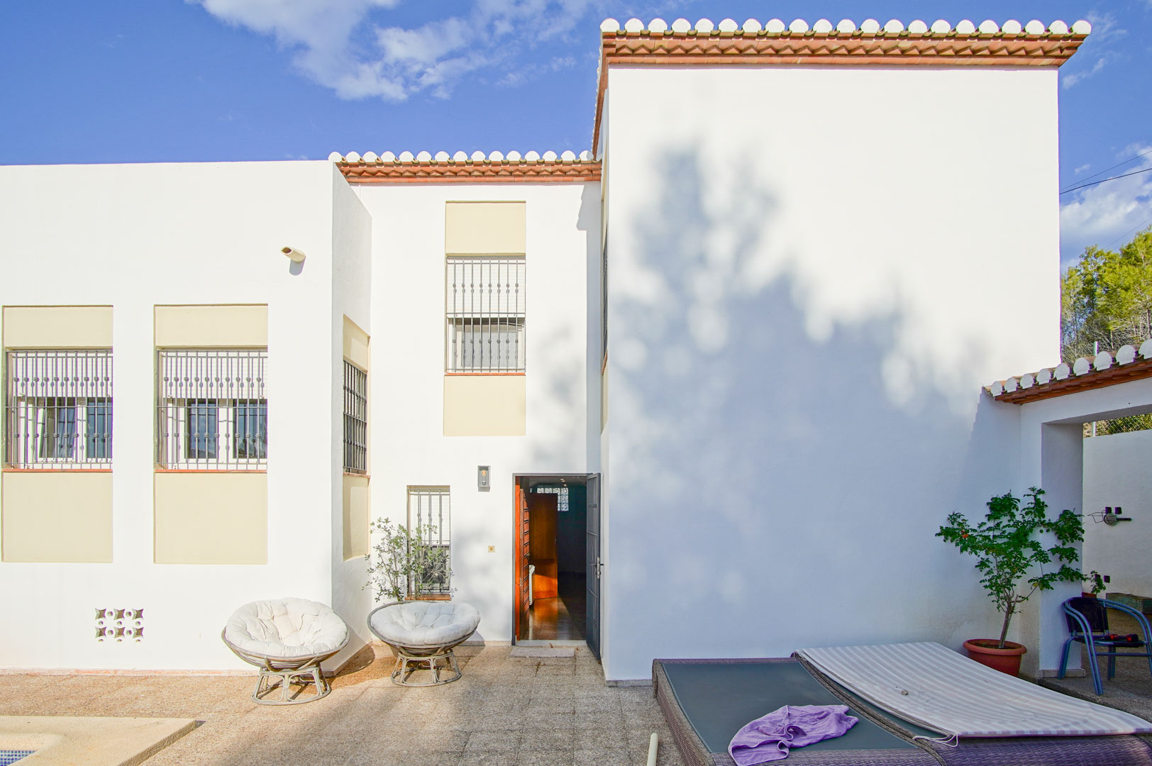 Fachada blanca de estilo mediterráneo con ventanas protegidas por rejas, terraza de piedra con mobiliario de exterior y acceso directo al interior de la vivienda.