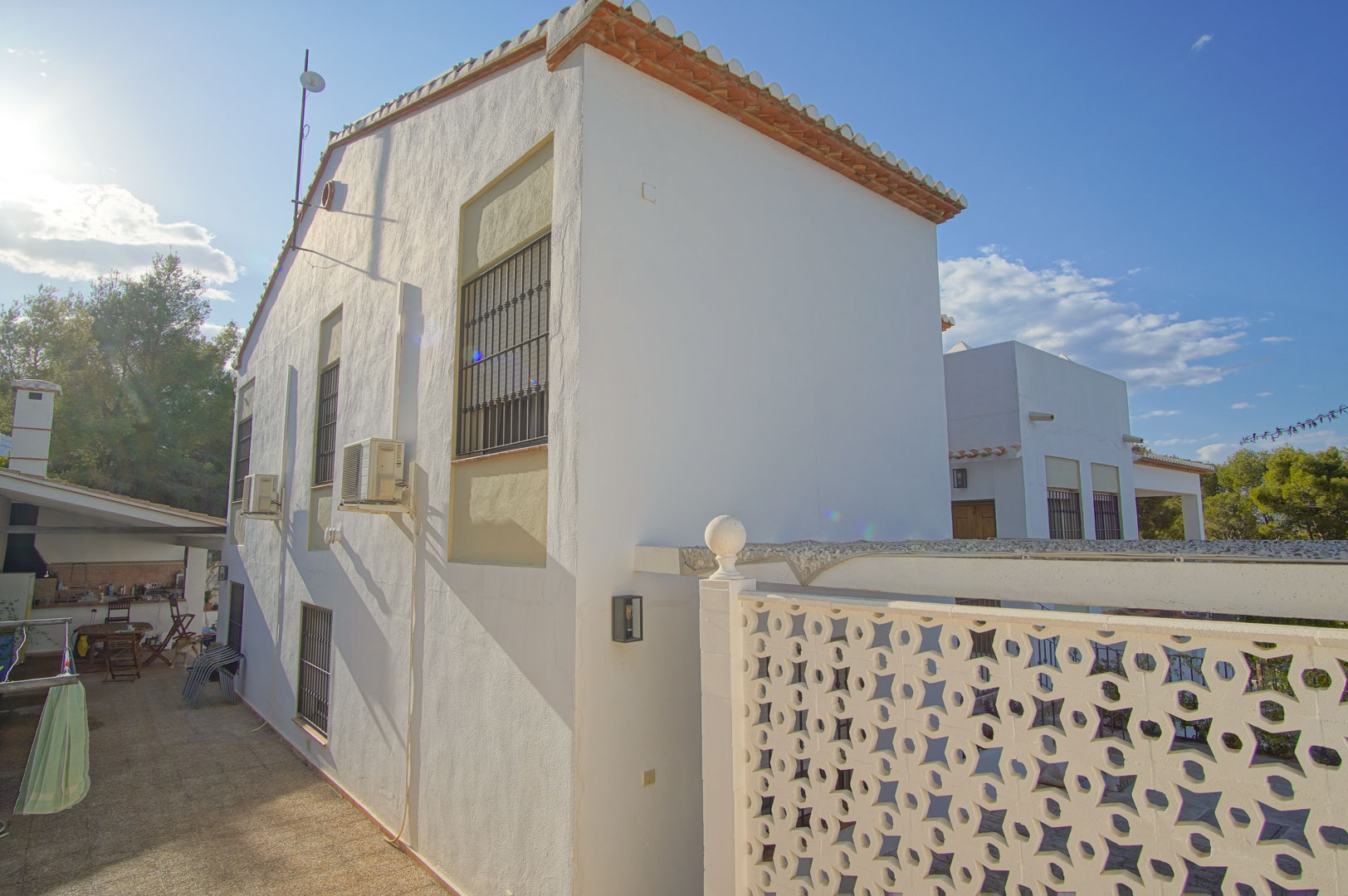 Fachada lateral de la villa con muros blancos, ventanas con rejas de seguridad y patio pavimentado que conduce a una zona de cocina exterior y barbacoa.