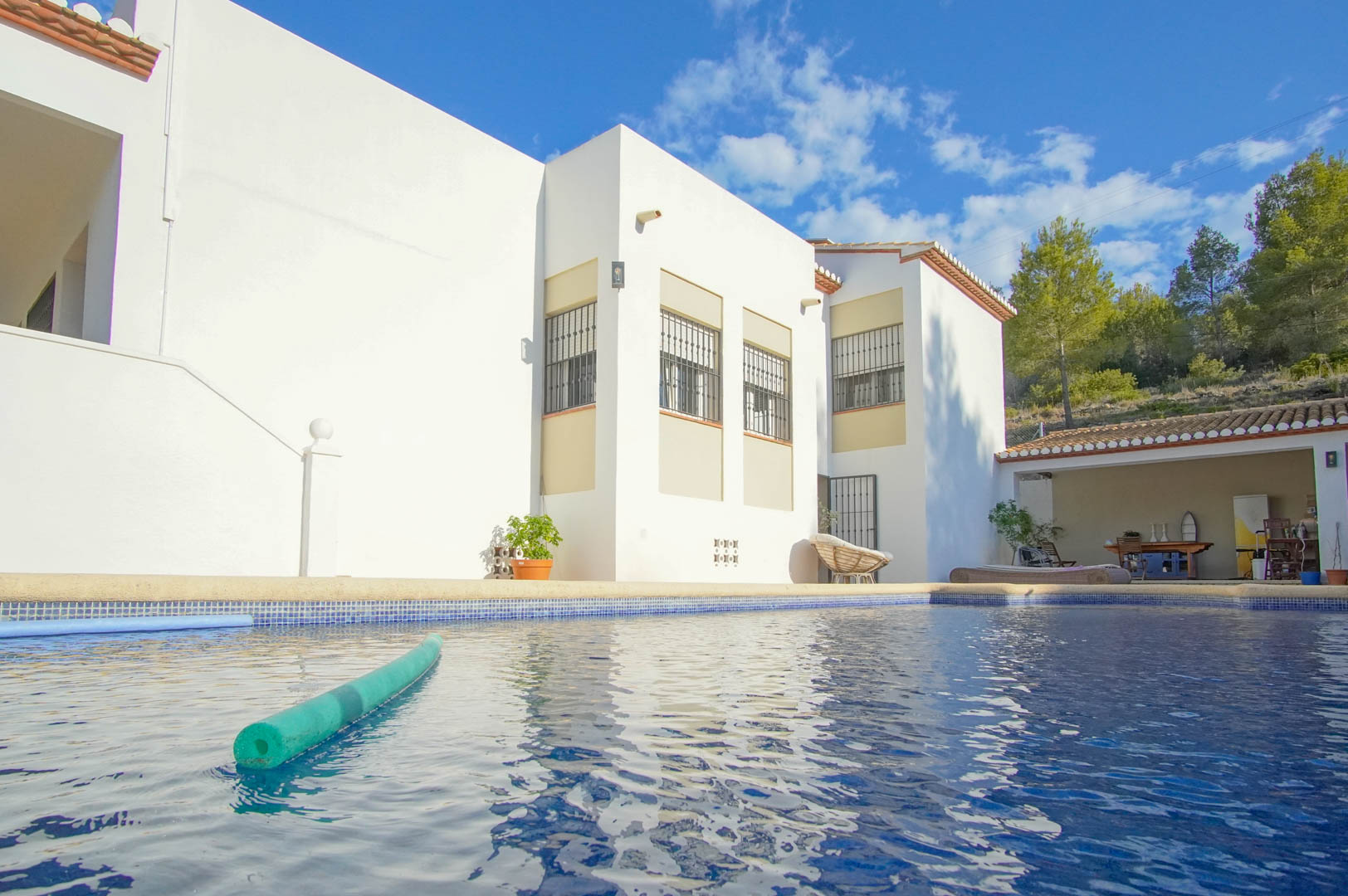 Piscina privada con revestimiento de gresite azul frente a la fachada blanca del chalet, que incluye una zona de porche cubierto y vistas al entorno natural.