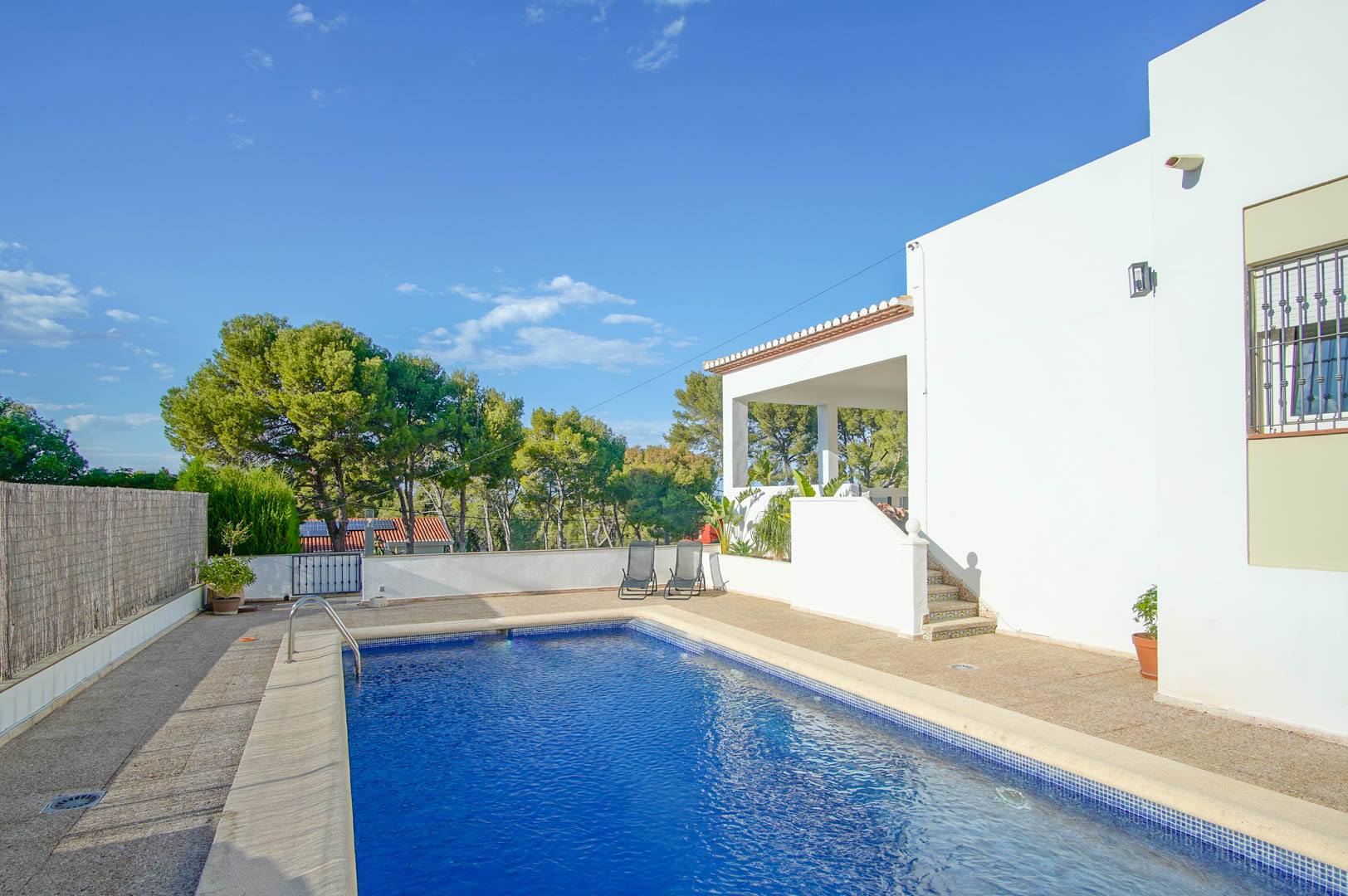 Piscina privada rectangular con revestimiento de gresite azul, rodeada de una amplia terraza de piedra y acceso directo a la planta principal de la villa blanca.