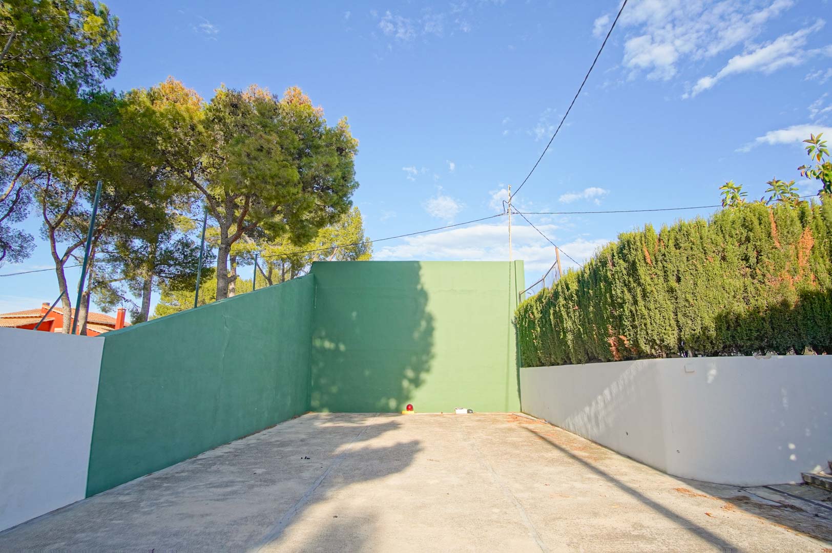 Pista de frontón privada con muros perimetrales en verde y suelo de hormigón, rodeada de vegetación madura y setos que garantizan privacidad.