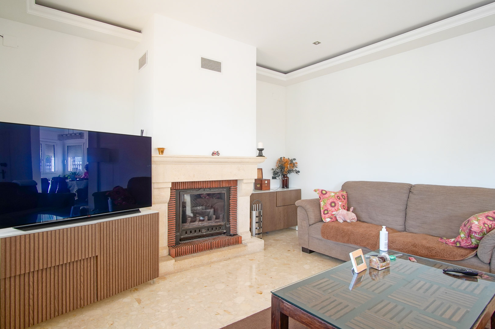 Salón luminoso con chimenea de piedra y ladrillo, suelos de mármol pulido y mobiliario de madera con listones verticales integrando la zona de televisión.