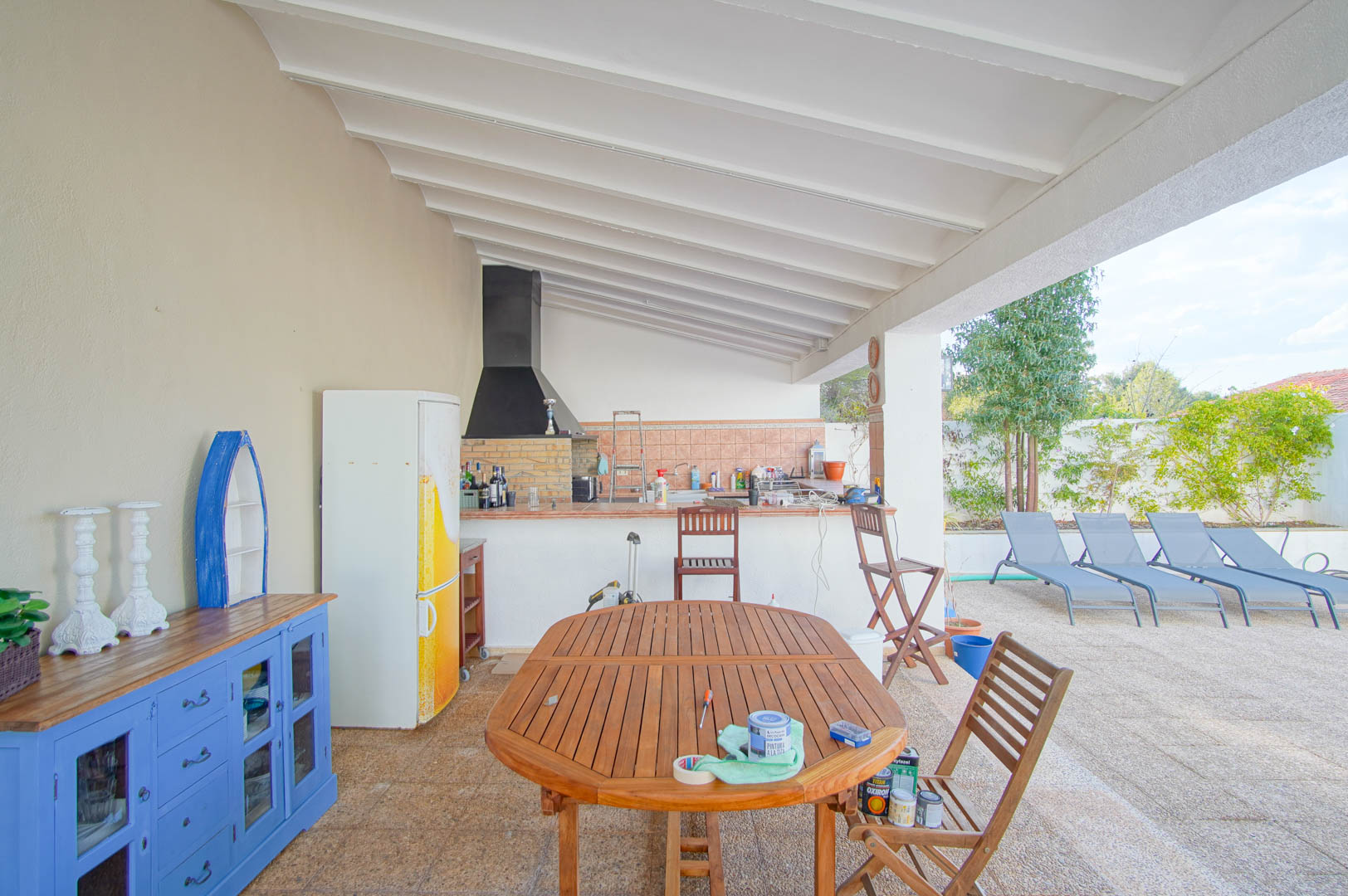 Terraza cubierta con cocina de verano, barbacoa de obra y techo de vigas blancas. Cuenta con comedor de madera y vistas directas a la zona de tumbonas junto a la piscina.
