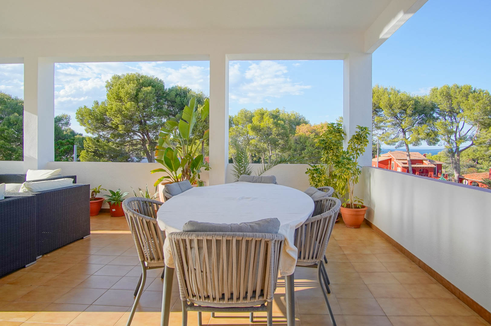 Terraza cubierta con comedor exterior y sofás de mimbre, ofreciendo vistas despejadas a la vegetación mediterránea y al mar en un entorno tranquilo.