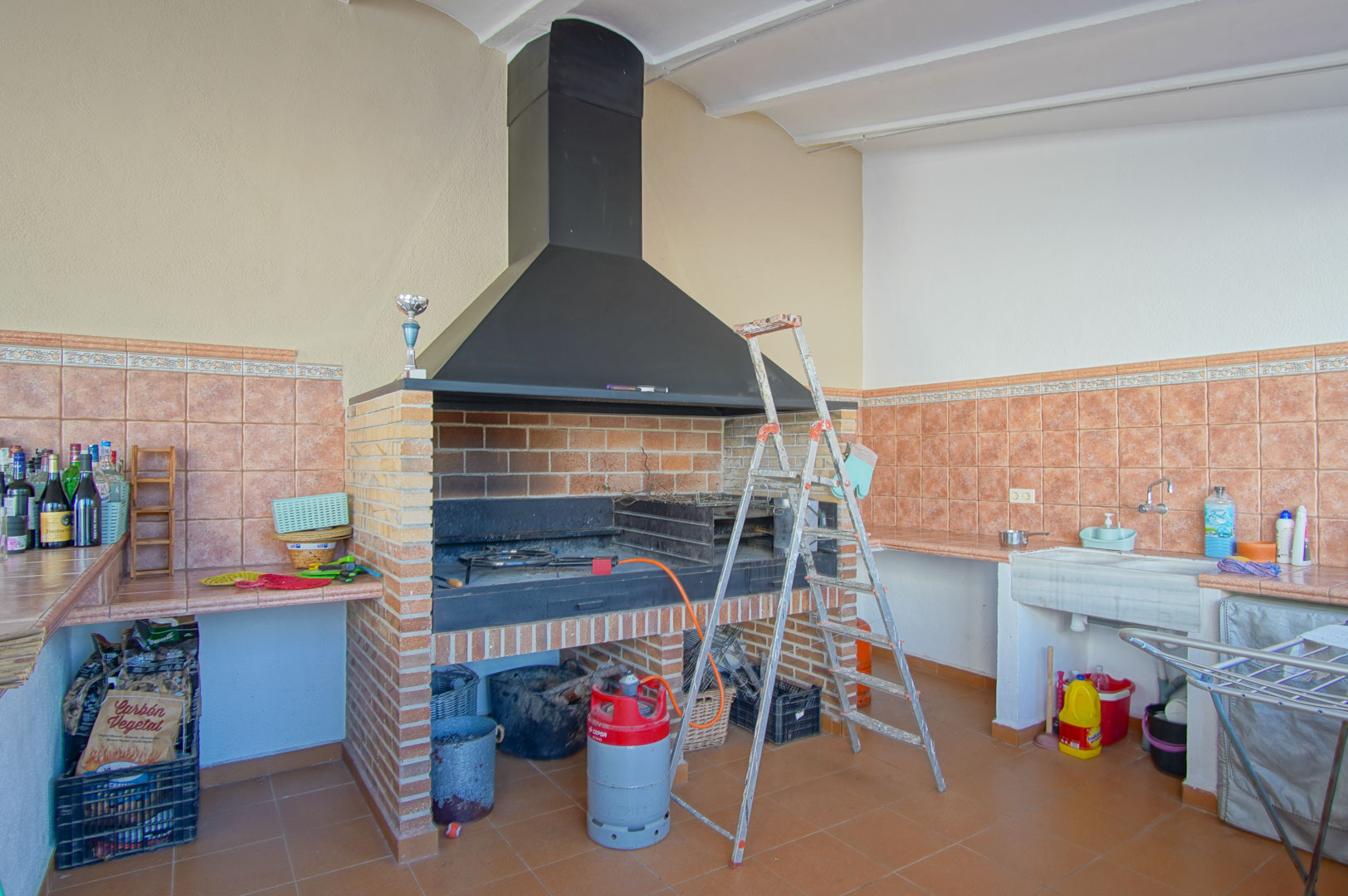 Zona de barbacoa de obra con campana de metal negro, fregadero de piedra y amplias encimeras alicatadas en tonos terracota para cocina exterior.