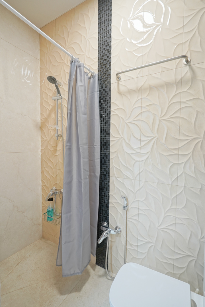 Baño con azulejos de relieve floral en tonos crema, acentuado por una franja vertical de mosaico negro y grifería cromada funcional.