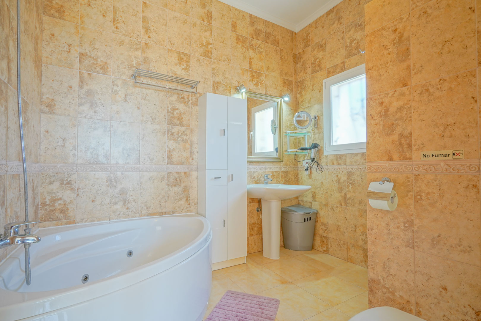 Cuarto de baño con bañera de hidromasaje rinconera, paredes revestidas en gres cerámico de tonos cálidos y mobiliario auxiliar blanco.