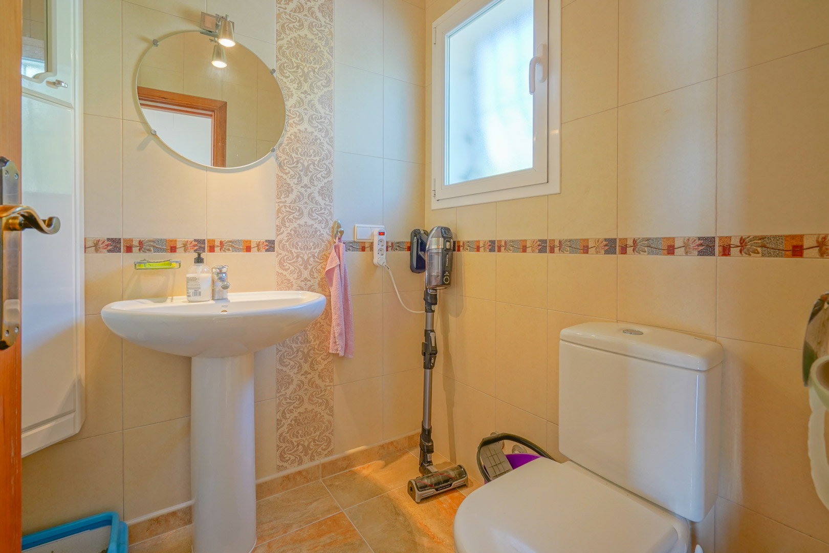 Cuarto de baño funcional con lavabo de pedestal, espejo circular y paredes alicatadas en tonos crema con cenefas decorativas y luz natural.