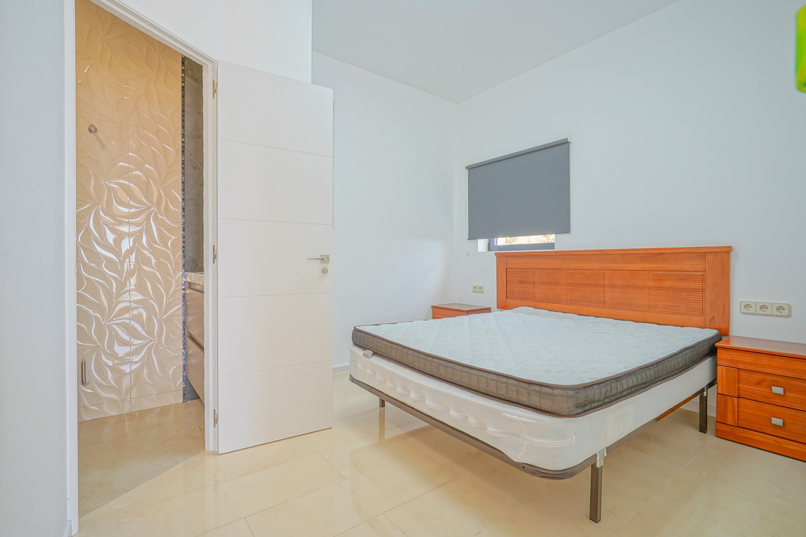 Dormitorio doble con mobiliario de madera, suelos cerámicos claros y acceso a un baño ensuite con azulejos decorativos de relieve ondulado.