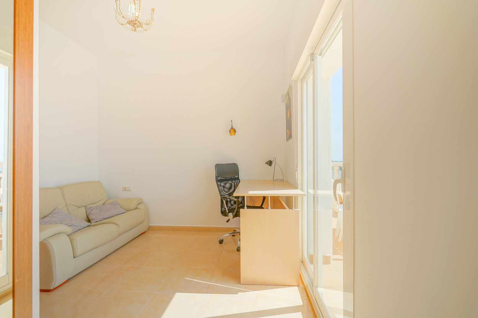 Habitación luminosa con zona de despacho, sofá de cuero beige y acceso directo a la terraza mediante ventanales. Suelos de gres en tonos cálidos y abundante luz natural.