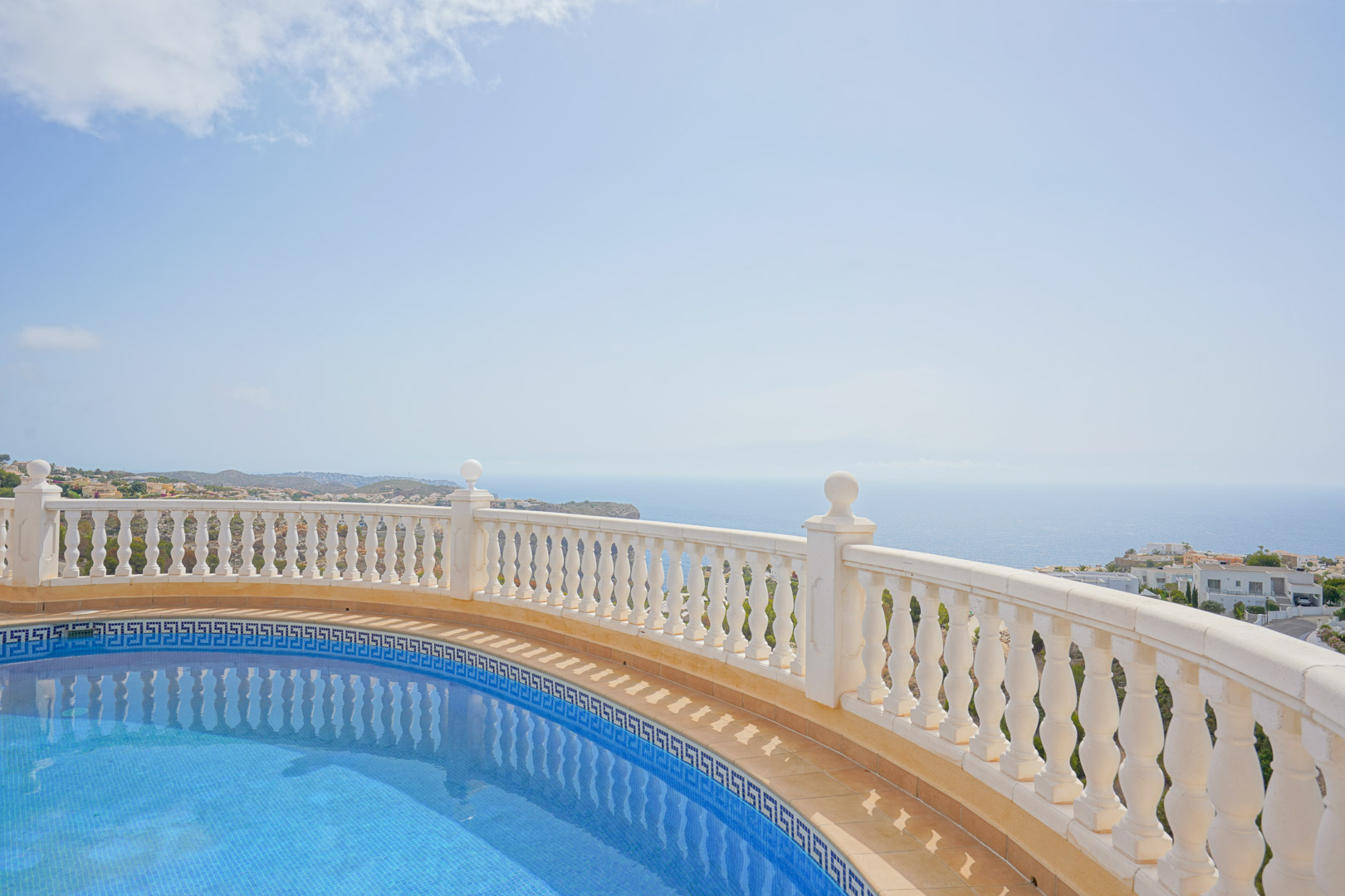 Piscina curva con mosaico azul y cenefa clásica, rodeada por una balaustrada blanca con vistas panorámicas al mar Mediterráneo y los acantilados.