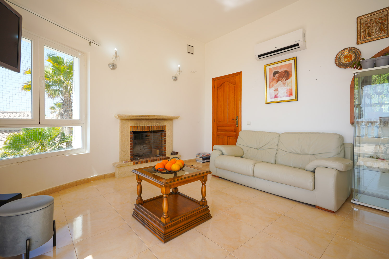 Salón con chimenea de piedra, sofá de piel beige y mesa de centro de madera. Incluye aire acondicionado y vistas exteriores a palmeras a través del ventanal.