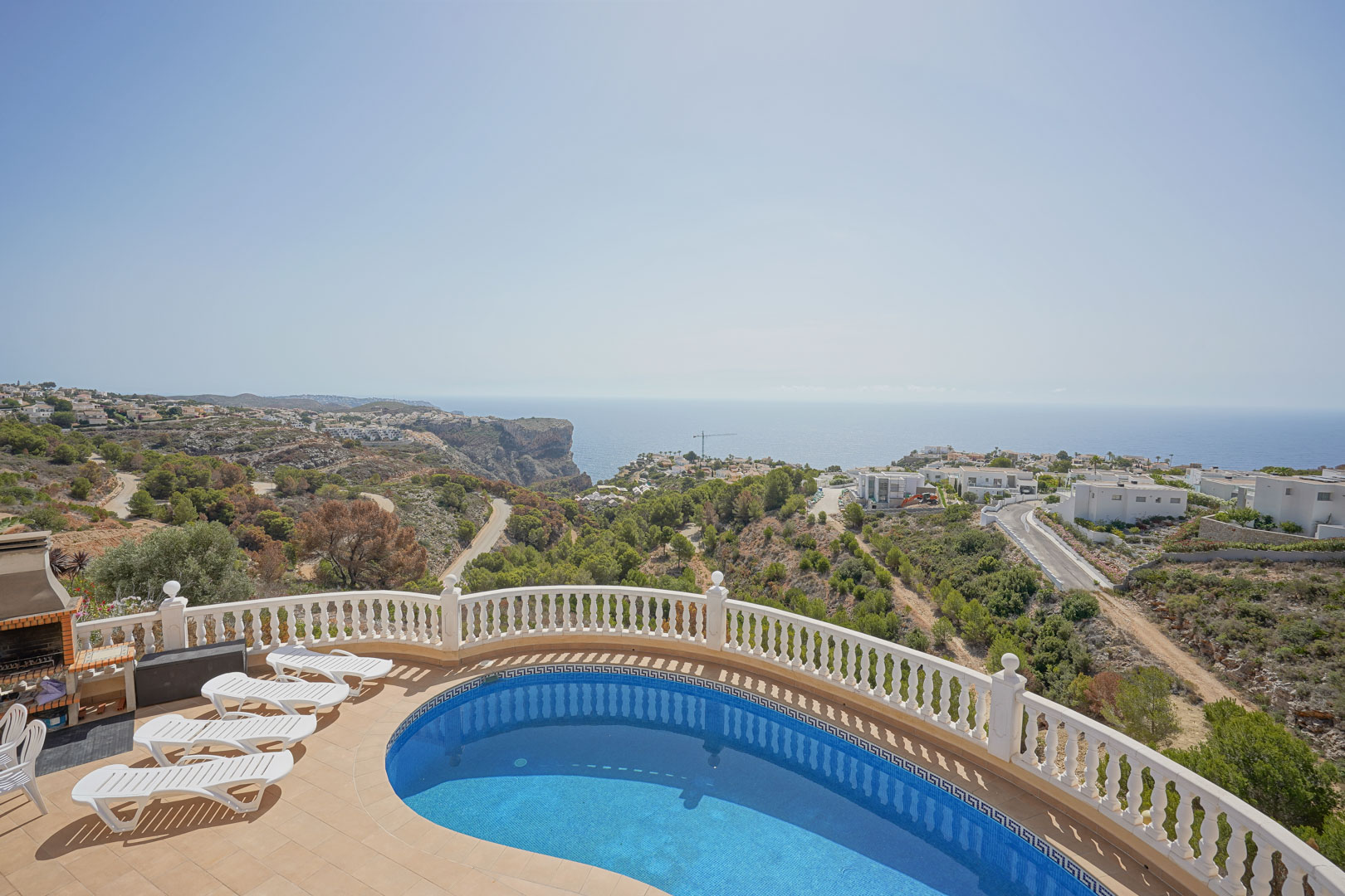 Terraza con piscina de forma riñonada, balaustrada blanca y zona de barbacoa de obra, ofreciendo vistas panorámicas al mar Mediterráneo y los acantilados de Cumbre del Sol.