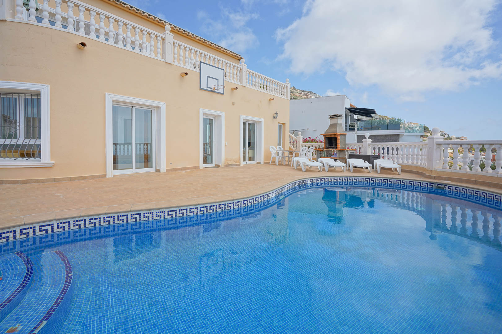 Terraza exterior con piscina de gresite azul y cenefa clásica. Incluye zona de barbacoa de obra, solárium con hamacas y fachada con canasta de baloncesto.