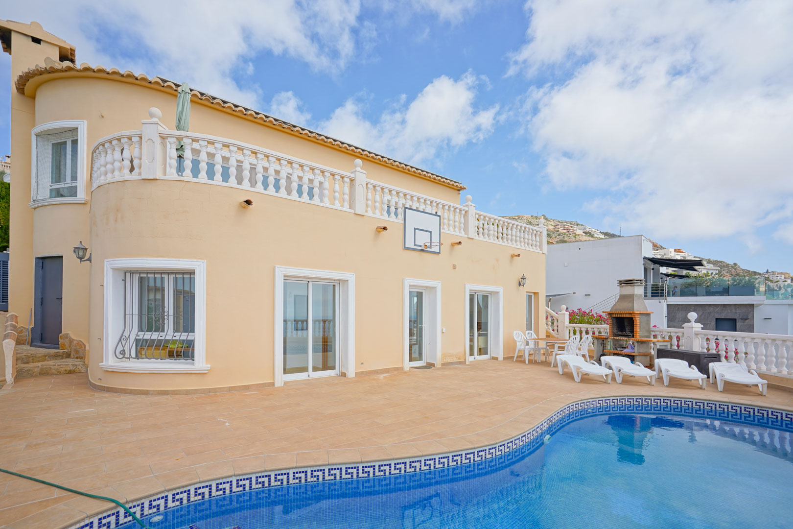 Terraza exterior con piscina de gresite azul, zona de barbacoa de obra y solárium. La fachada curva integra ventanales blancos y una balaustrada clásica en la planta superior.