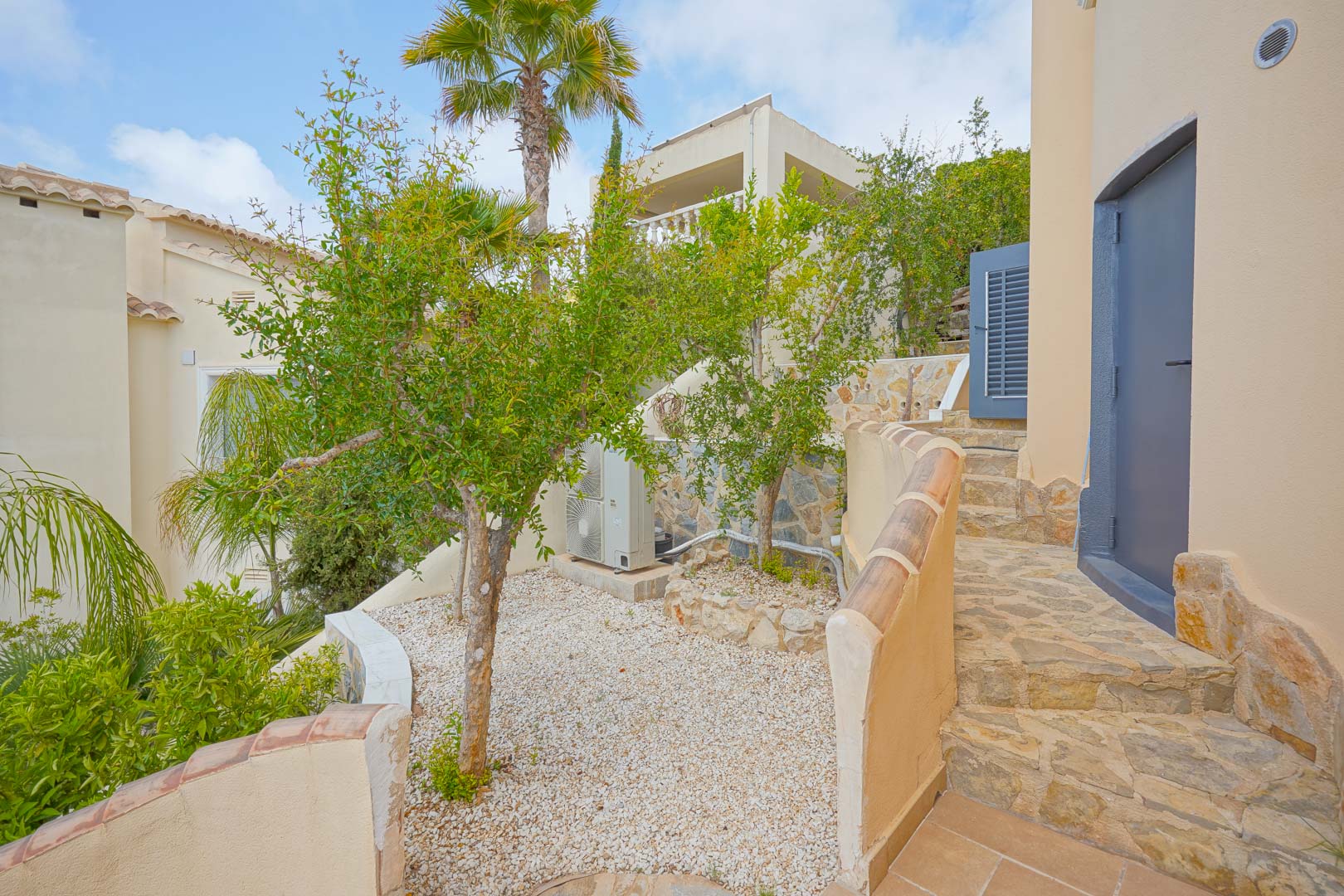 Terraza lateral con árboles frutales, pavimentada con piedra natural y grava blanca, comunicada mediante escaleras exteriores de piedra.