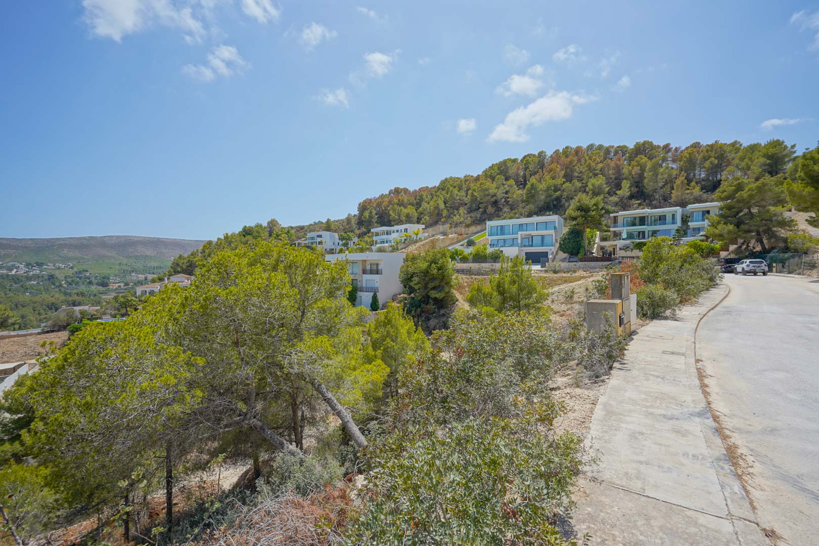 Calle pavimentada en una urbanización moderna con villas contemporáneas integradas en la ladera arbolada y aceras de hormigón.