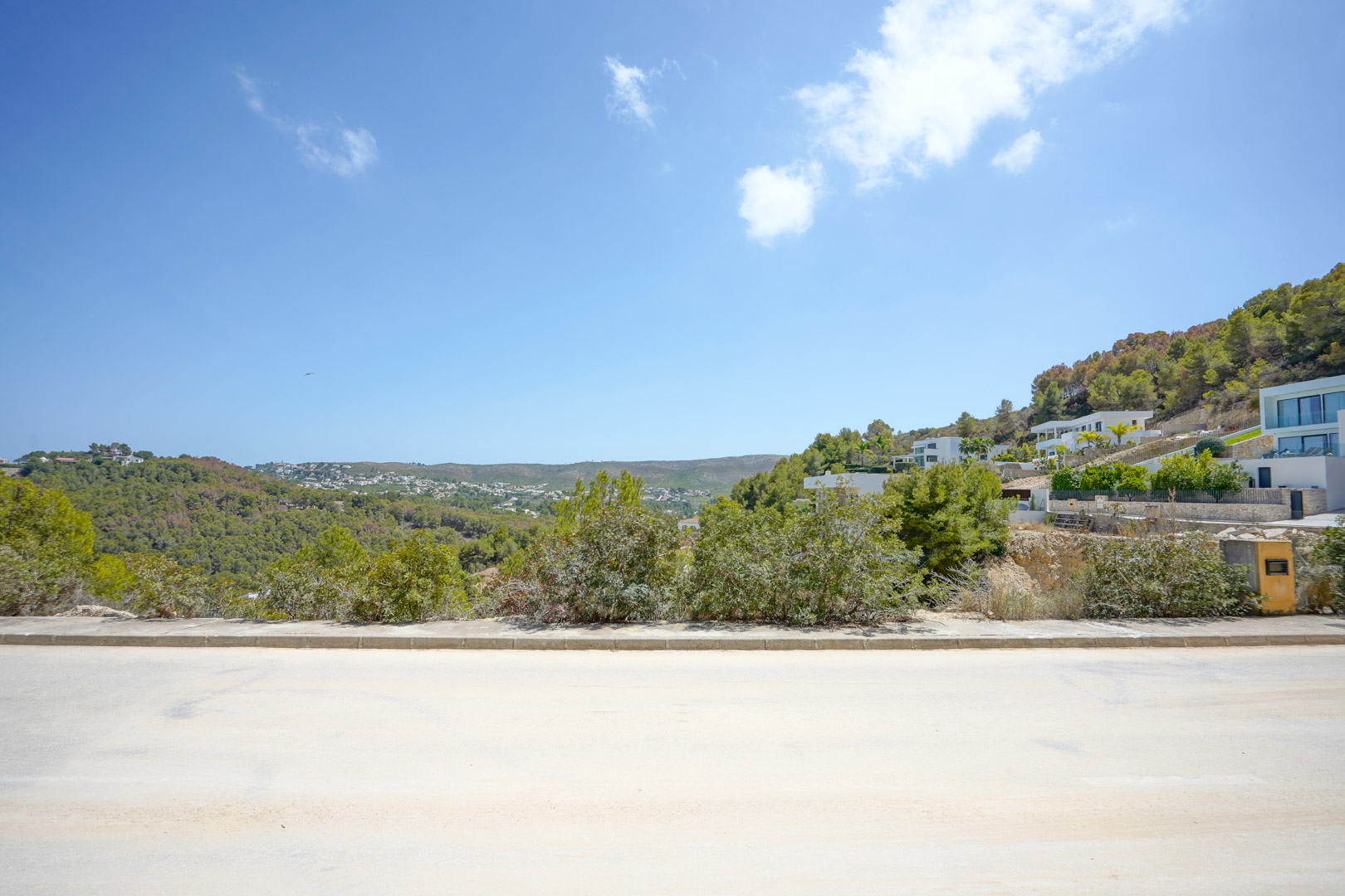Vista de la calle asfaltada con aceras en una urbanización moderna, mostrando la elevación del terreno y las colinas circundantes bajo un cielo despejado.