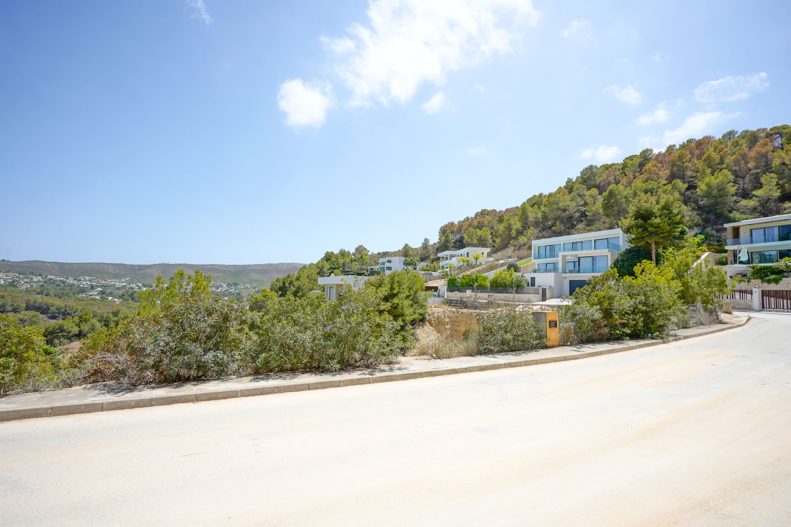 Vista de la calle pavimentada en una urbanización moderna, mostrando villas de arquitectura contemporánea integradas en la ladera boscosa con vistas despejadas.