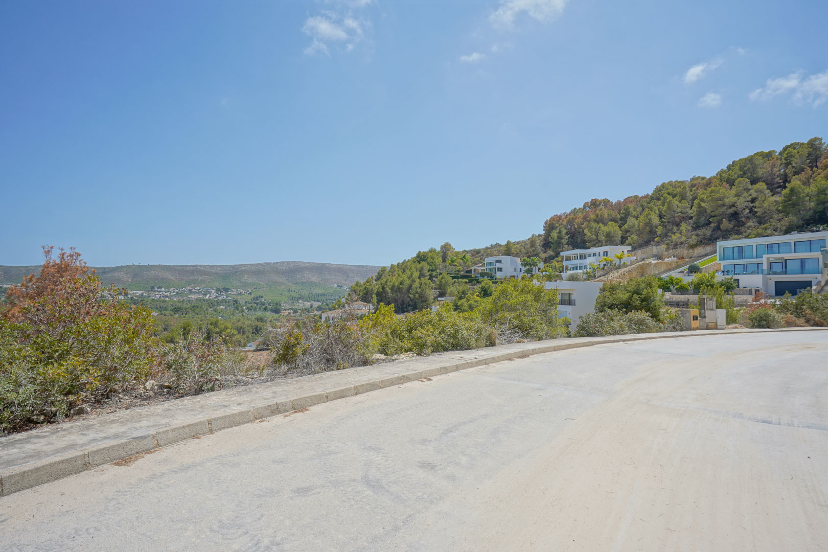 Vista desde el vial de acceso pavimentado que muestra la topografía inclinada y las villas modernas integradas en la colina boscosa bajo un cielo despejado.