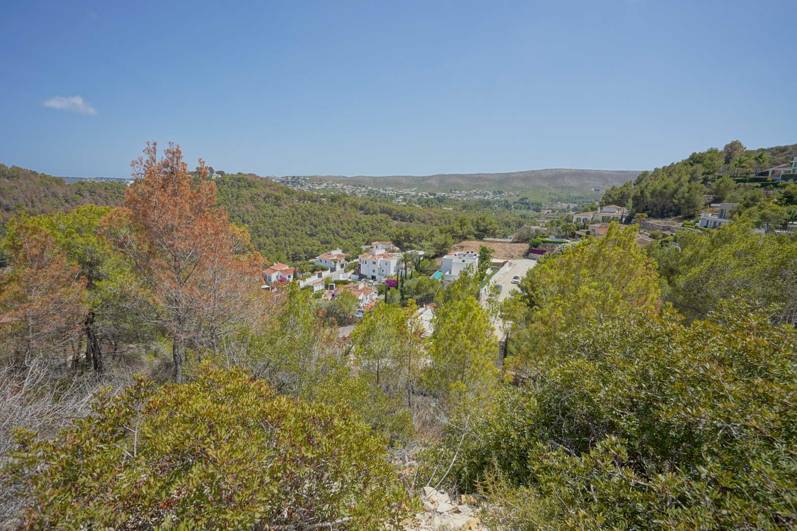 Vista elevada desde la parcela hacia el valle, mostrando la vegetación mediterránea autóctona y villas integradas en el paisaje montañoso bajo un cielo despejado.