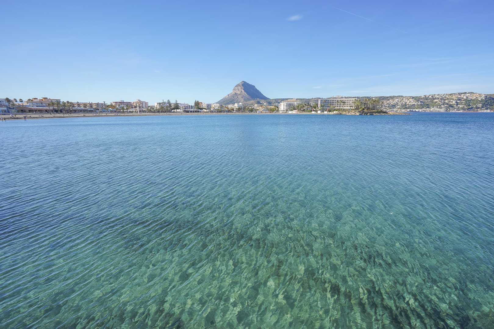 Vista panorámica de la bahía con aguas cristalinas color turquesa, la playa de arena y el perfil montañoso al fondo bajo un cielo despejado.