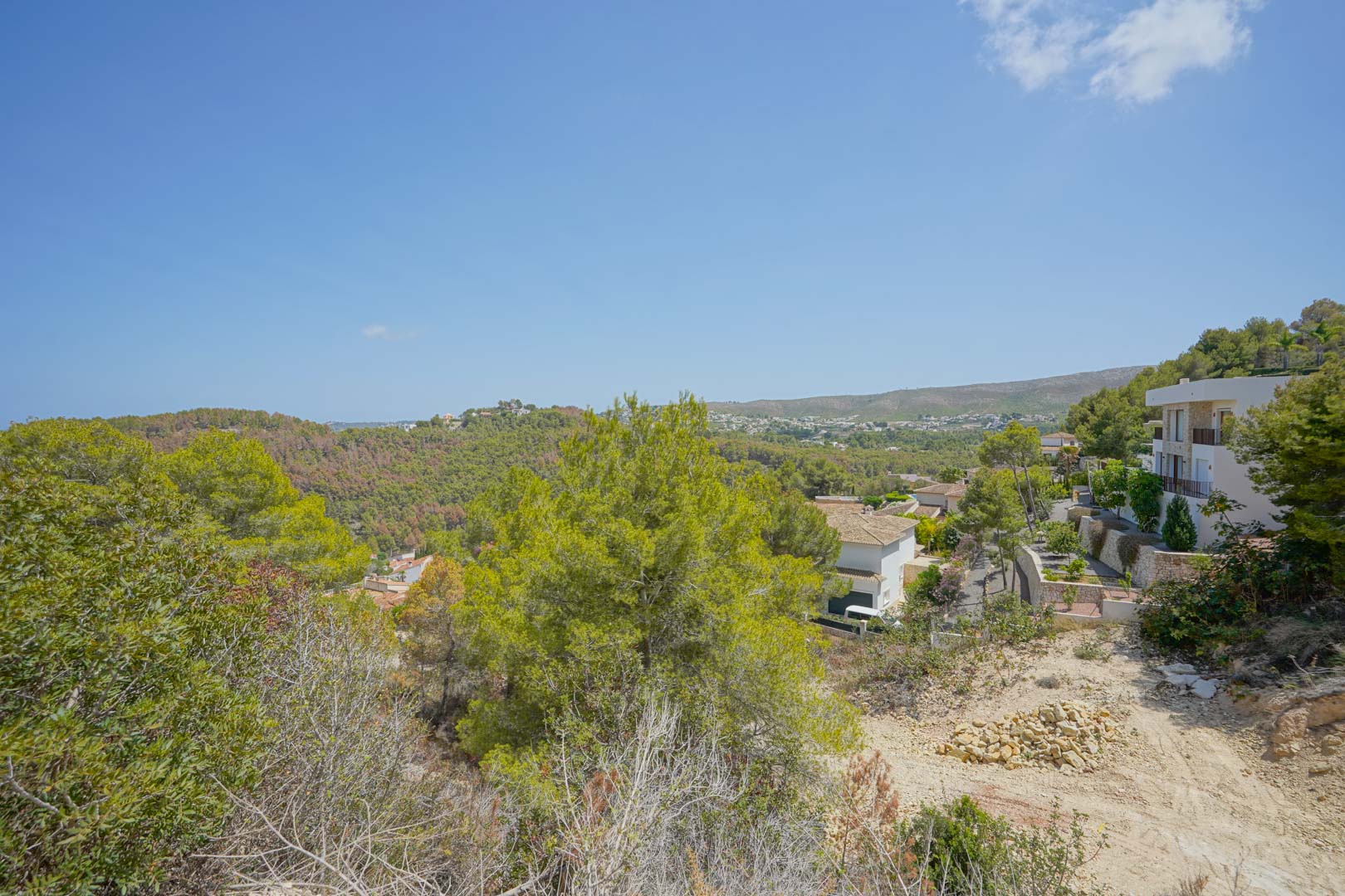 Vista panorámica desde la parcela elevada que muestra el terreno natural, la vegetación autóctona y las villas circundantes con vistas a las colinas y al mar.