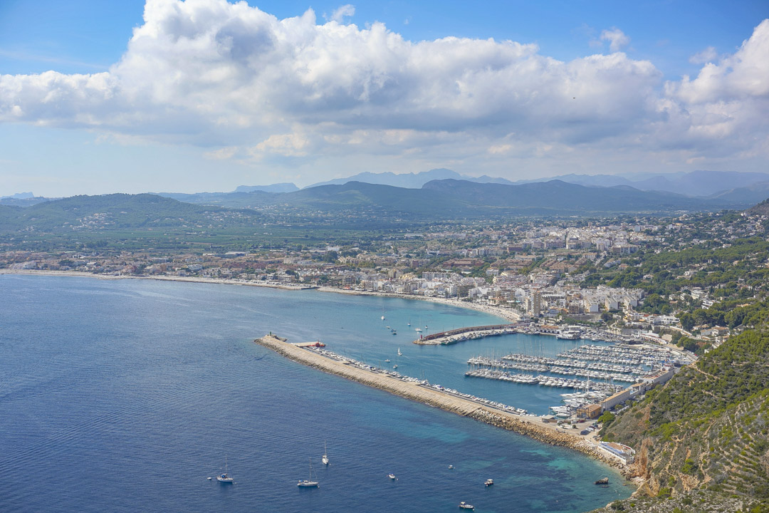 Vista panorámica elevada del puerto deportivo y la costa, mostrando el rompeolas, las embarcaciones de recreo y las montañas al fondo bajo un cielo despejado.