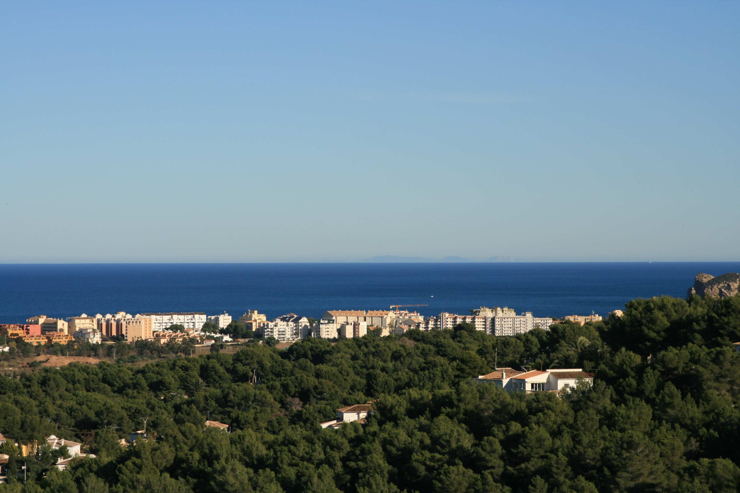 Vista panorámica elevada que muestra un extenso pinar en primer plano, seguido por una zona urbana costera y el mar Mediterráneo bajo un cielo despejado.