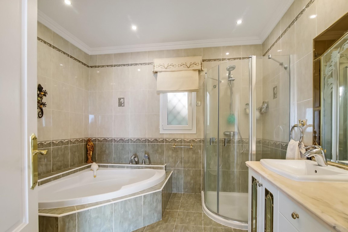 Baño completo con bañera de rincón, cabina de ducha curva y mueble de lavabo con encimera de mármol. Revestimiento cerámico con cenefas decorativas e iluminación empotrada.