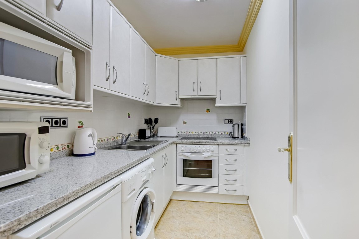 Cocina funcional con armarios blancos, encimera de granito gris y electrodomésticos integrados. Incluye zona de lavandería y molduras decorativas en el techo.