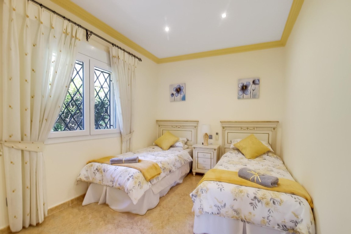Dormitorio con dos camas individuales, cabeceros clásicos blancos, iluminación empotrada y ventana con rejas de seguridad y cortinas claras.