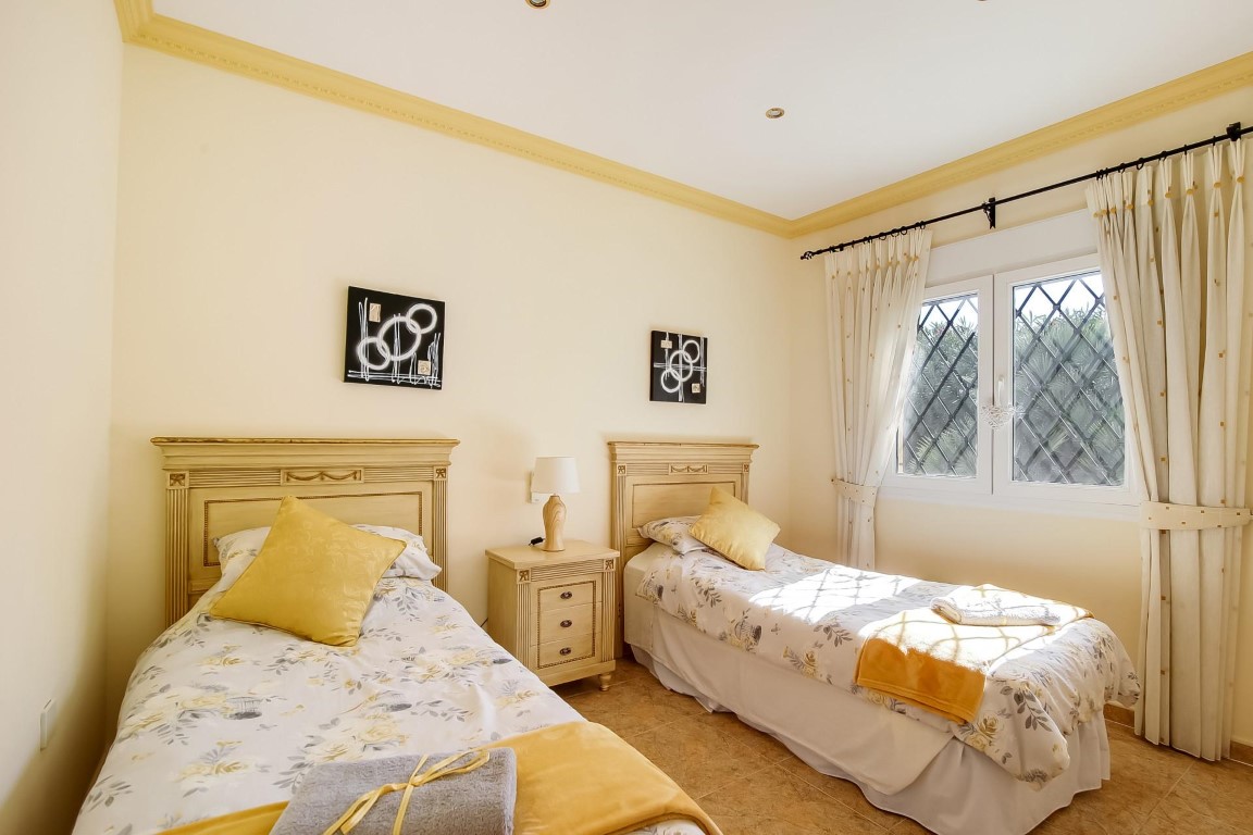 Dormitorio con dos camas individuales de estilo clásico, cabeceros de madera, molduras decorativas en el techo y ventana con rejería de seguridad en forma de rombos.