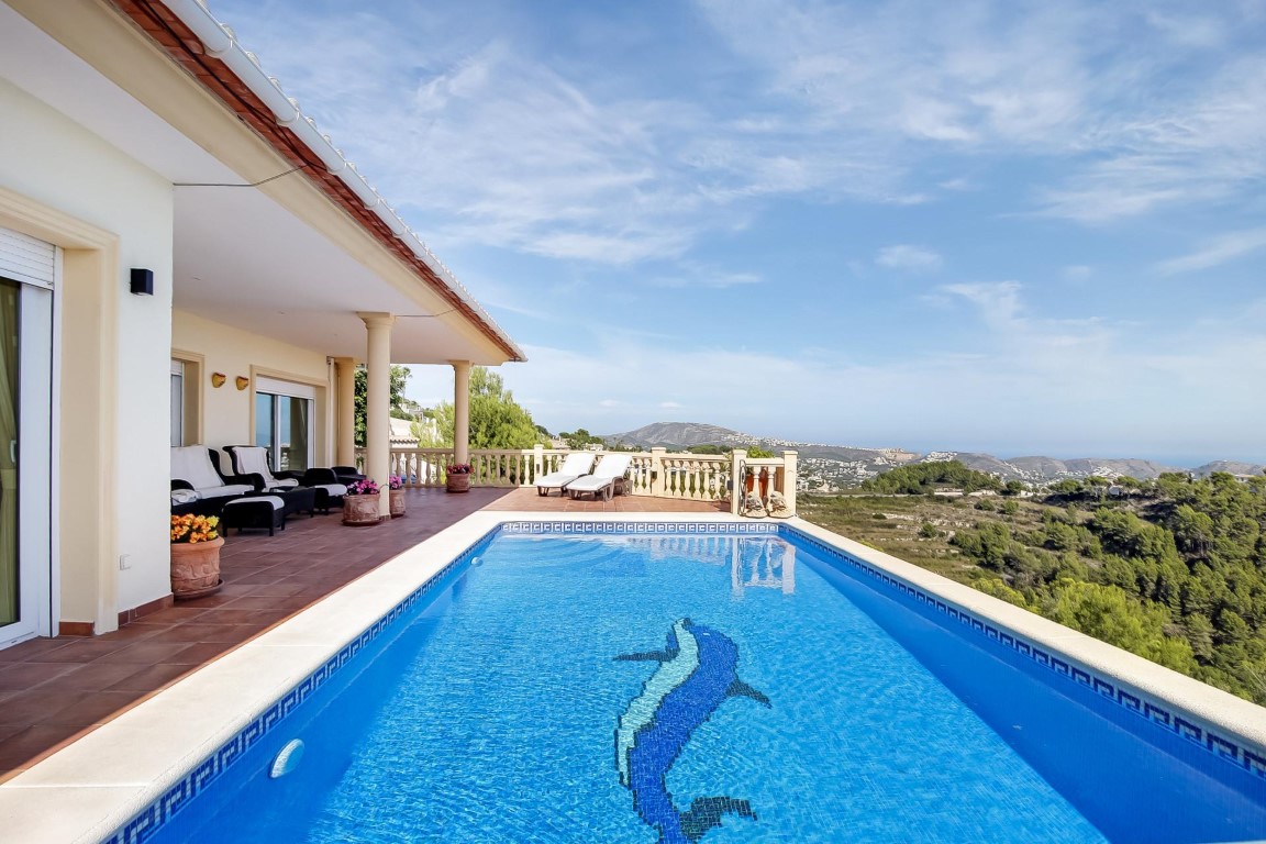 Piscina exterior con mosaico de delfín y amplia terraza de terracota, ofreciendo vistas panorámicas al mar y al valle de Moraira desde la zona de porche.
