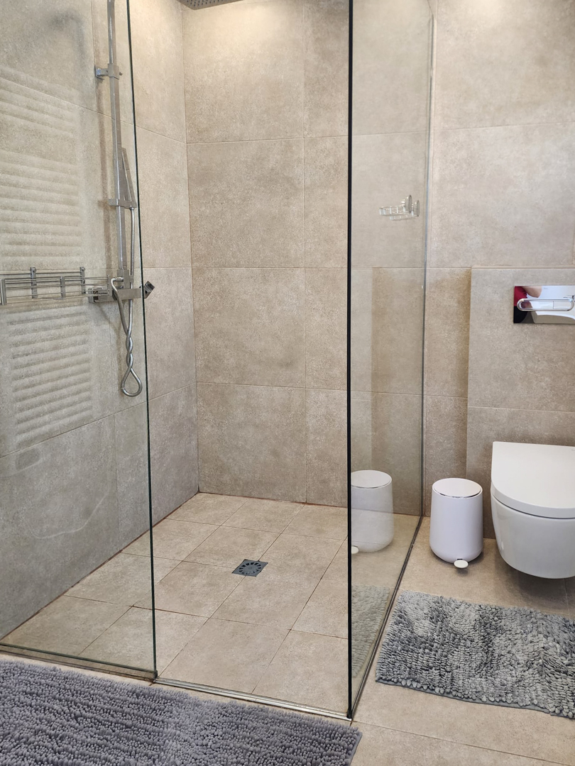 Baño moderno con ducha a ras de suelo, mampara de cristal fija y azulejos porcelánicos en tonos arena. Incluye inodoro suspendido y grifería cromada de diseño contemporáneo.