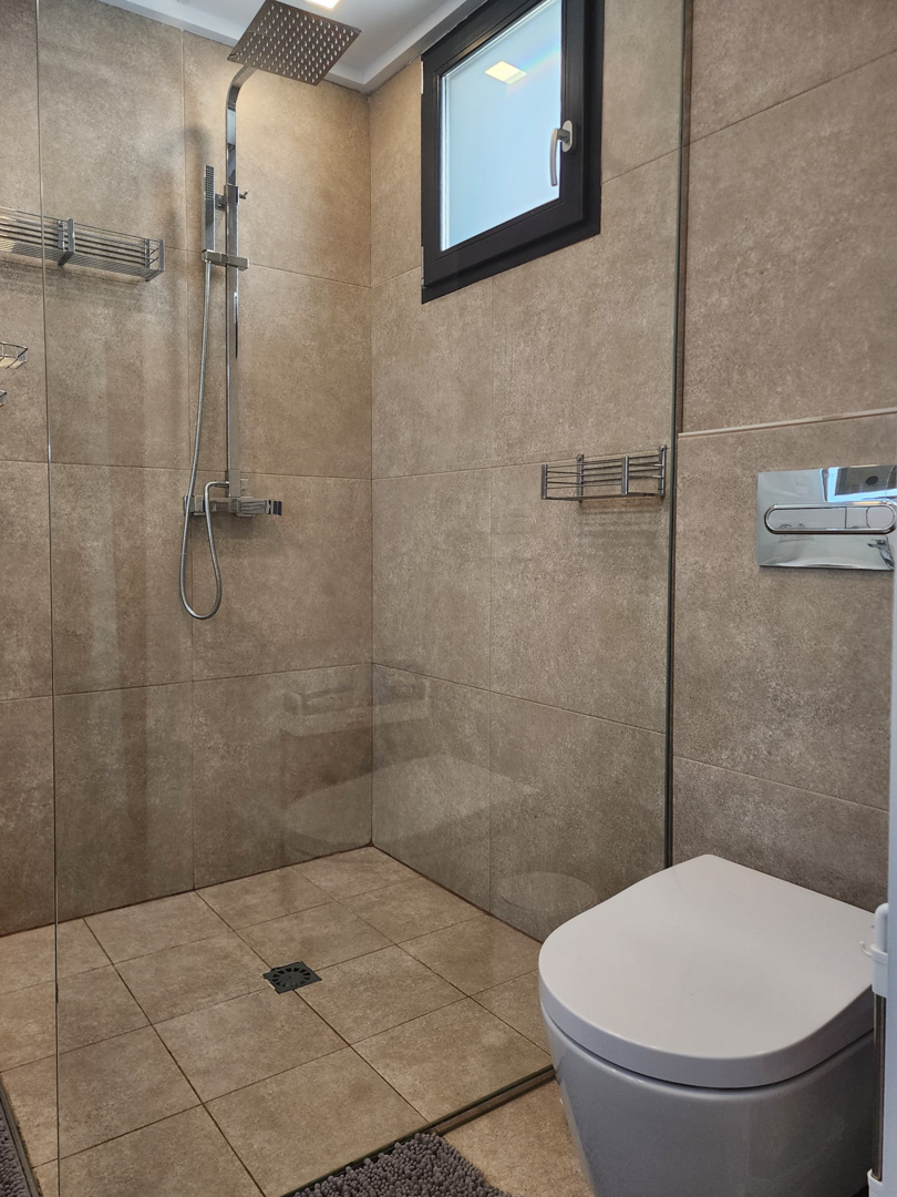 Baño moderno con ducha a ras de suelo, mampara de cristal, grifería cromada de efecto lluvia y revestimiento cerámico integral en tonos arena naturales.