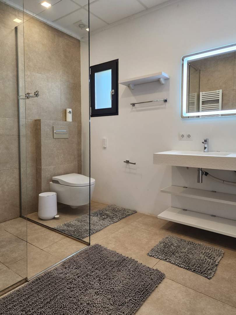 Baño moderno con ducha a ras de suelo, mampara de cristal, inodoro suspendido y lavabo con estantes abiertos y espejo retroiluminado LED sobre azulejos beige.