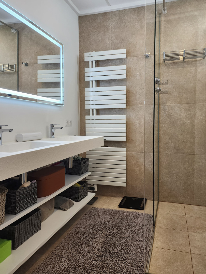 Baño moderno con lavabo doble integrado, estanterías abiertas y espejo con iluminación LED. Incluye radiador toallero blanco y ducha de obra con azulejos en tonos tierra.