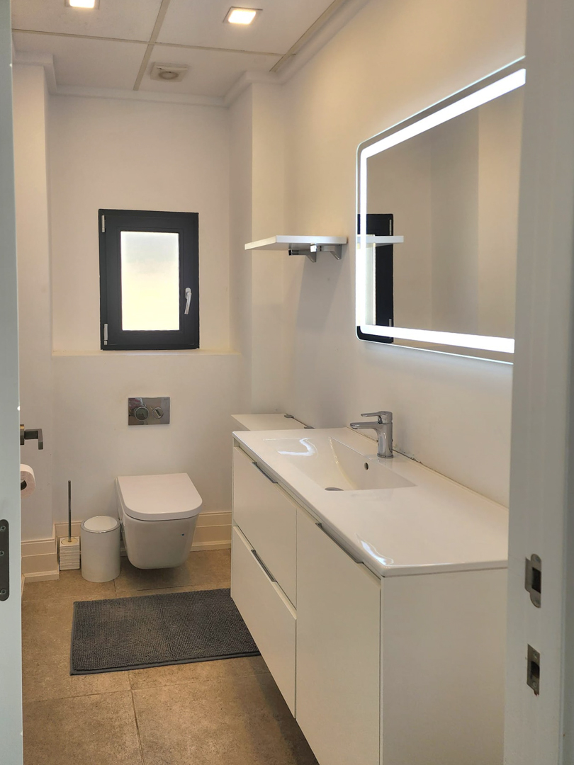 Baño moderno con mueble de lavabo blanco, espejo retroiluminado por LED y sanitario suspendido. Presenta suelos cerámicos en tonos neutros y ventana de ventilación.