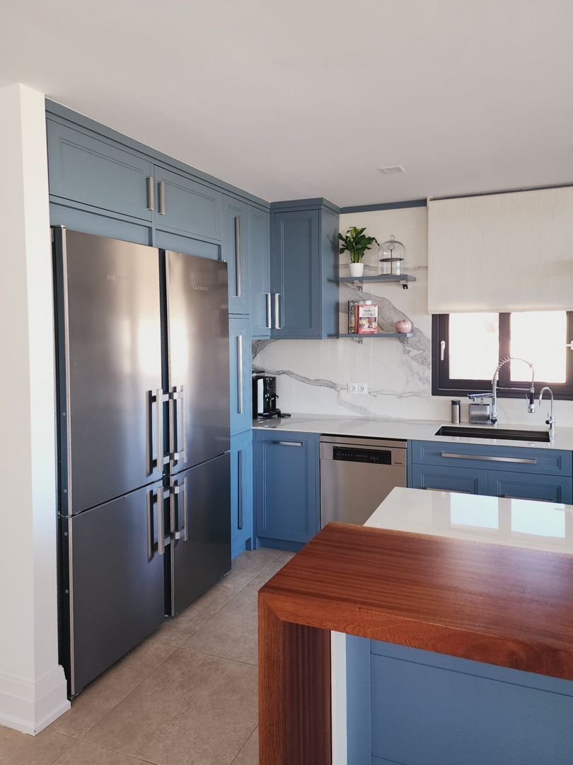 Cocina de diseño con armarios en azul empolvado, encimera blanca y salpicadero de mármol. Incluye electrodomésticos de alta gama y una barra de madera natural.