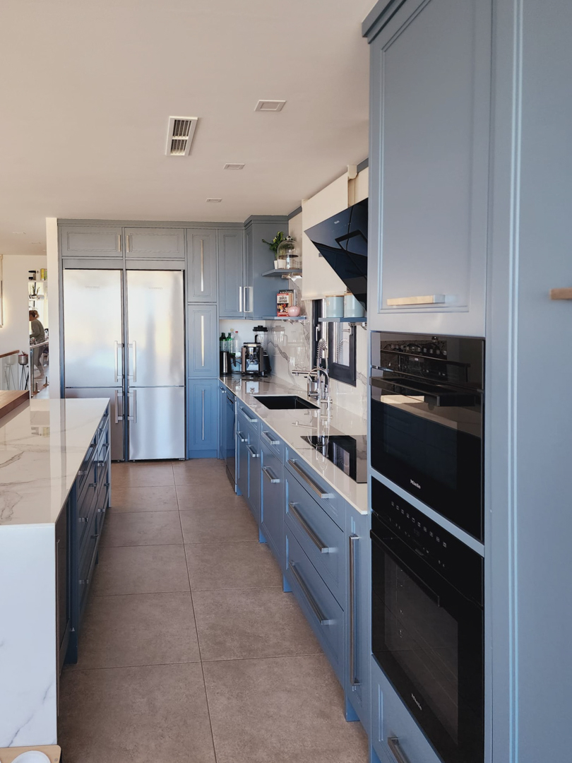 Cocina de diseño con mobiliario azul grisáceo, encimeras de efecto mármol y electrodomésticos Miele integrados, incluyendo hornos dobles y un gran frigorífico de acero inoxidable.
