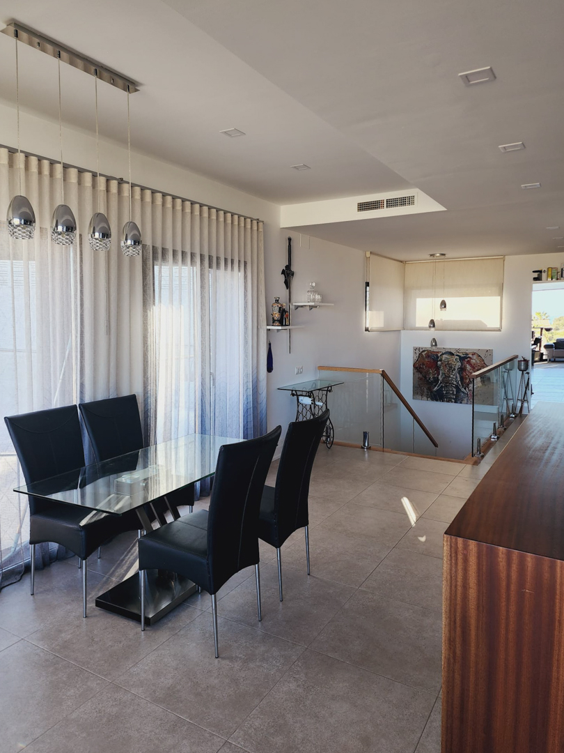 Comedor con mesa de cristal y sillas negras, junto a una escalera con barandilla de vidrio. Destacan las lámparas colgantes cromadas y el suelo de gres porcelánico.