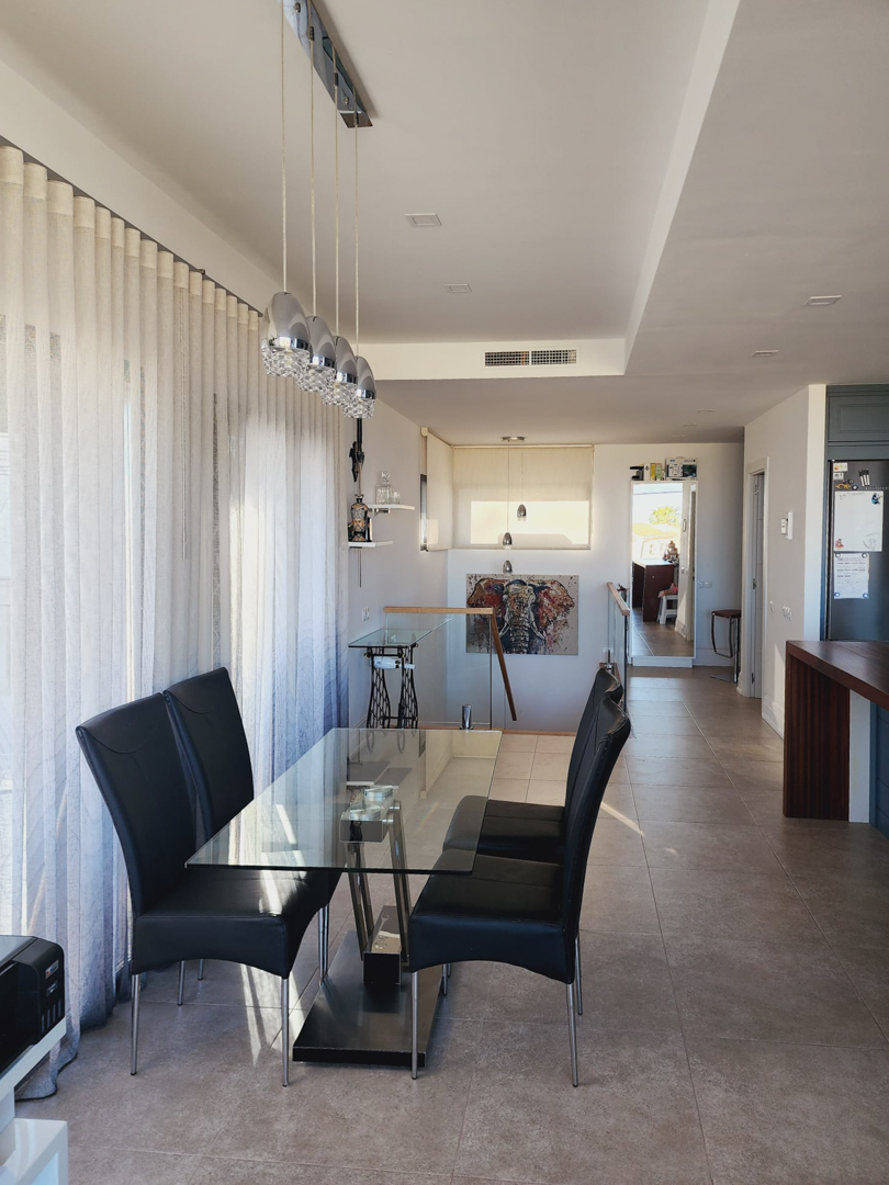 Comedor luminoso con mesa de cristal, sillas de cuero negro y lámparas colgantes de diseño, junto a ventanales con cortinas traslúcidas y acceso a la planta inferior.