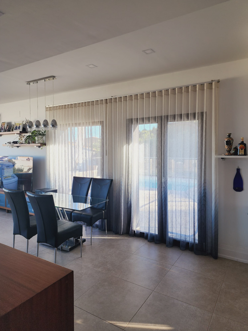 Comedor luminoso con mesa de cristal y sillas de respaldo alto, junto a ventanales con cortinas degradadas que dan acceso directo a la zona exterior de la piscina.