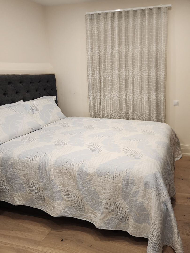 Dormitorio moderno con cama doble, cabecero capitoné en gris oscuro, suelos de madera clara y cortinas de techo a suelo con diseño geométrico.