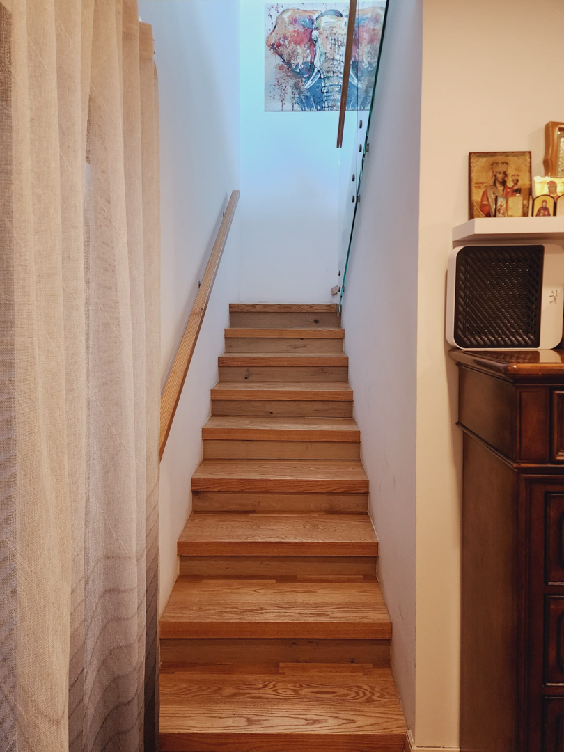 Escalera interior de madera con pasamanos a juego y barandilla de cristal, iluminada por luz natural superior y decorada con elementos artísticos.